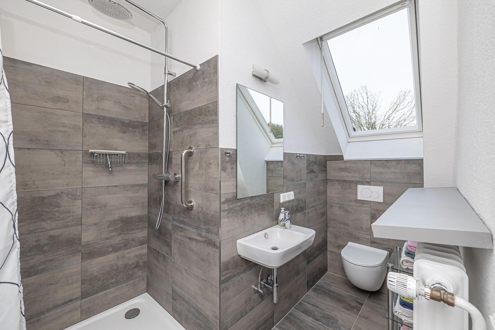 Badezimmer mit Dusche, Waschbecken und WC. Dachfenster und graue Fliesen.