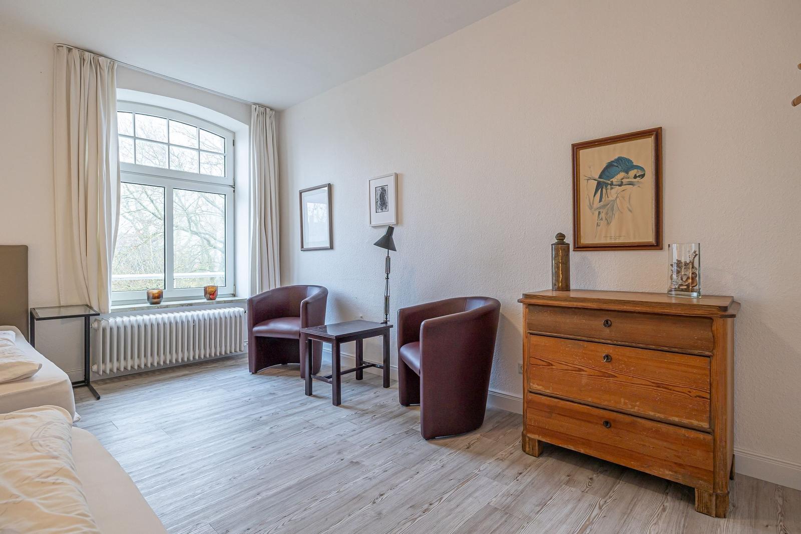 Ein Zimmer mit zwei Sesseln, einem Schrank und einem Fenster mit Blick auf Bäume.