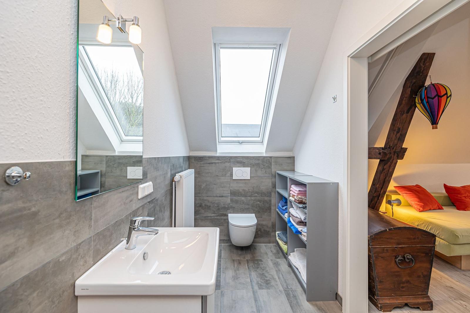 Badezimmer mit Waschbecken, Toilette und Dachfenster. Nebenraum mit Bett und Holzkonstruktion.