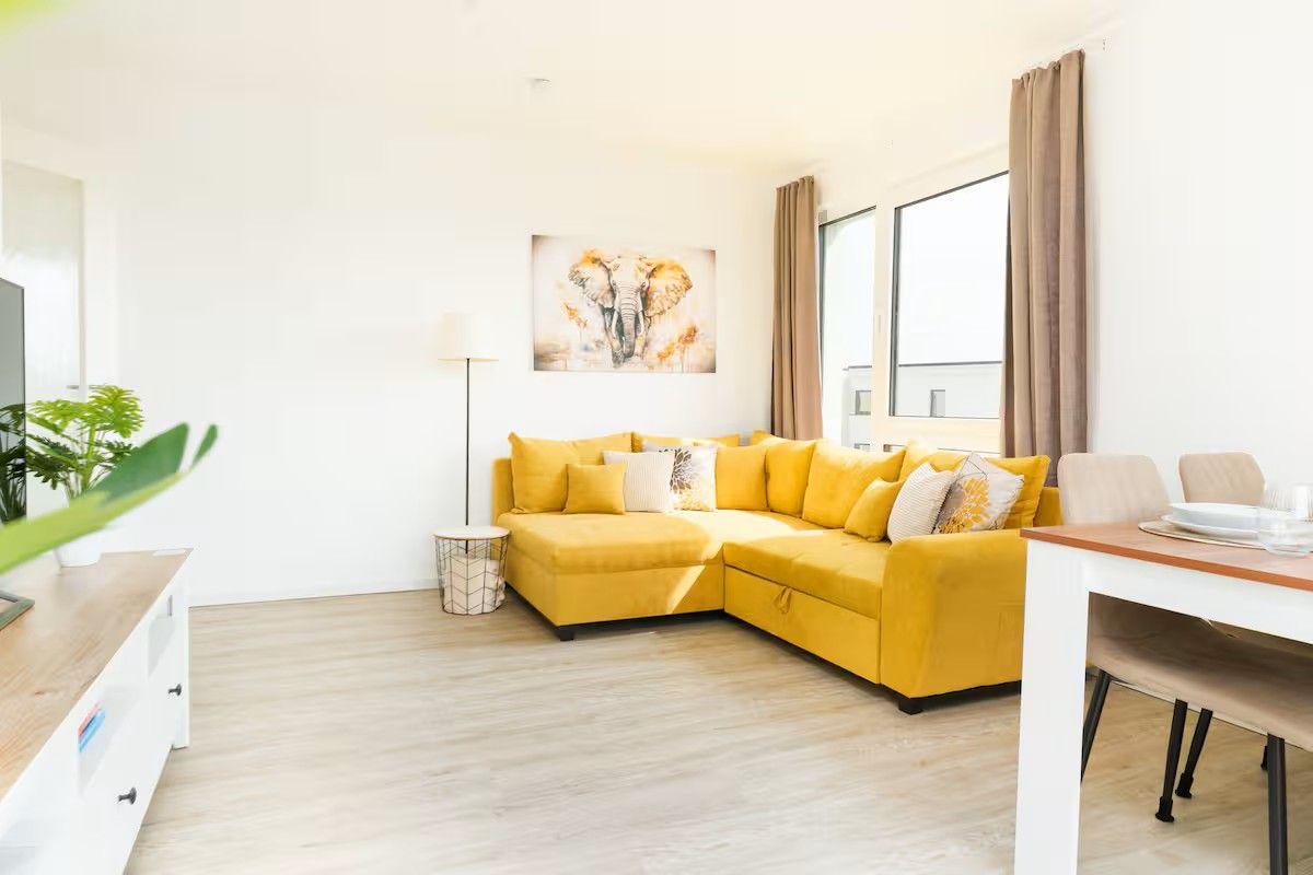 Ein Wohnzimmer mit gelbem Ecksofa, Wandbild und Essbereich.