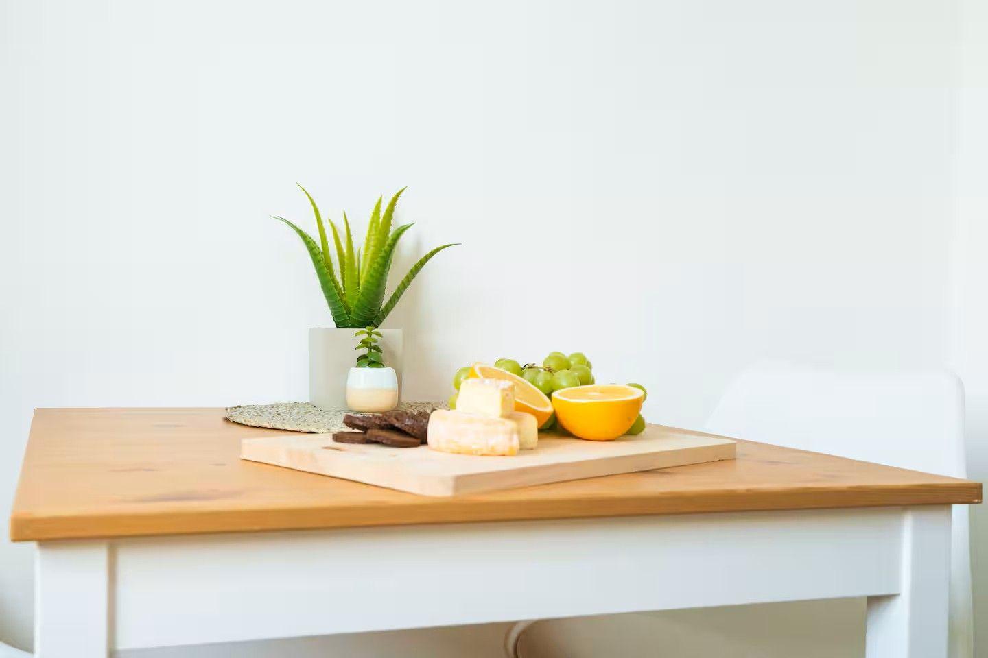 Holztisch mit Käse, Obst und Pflanze. Weiße Stühle und Wand im Hintergrund.