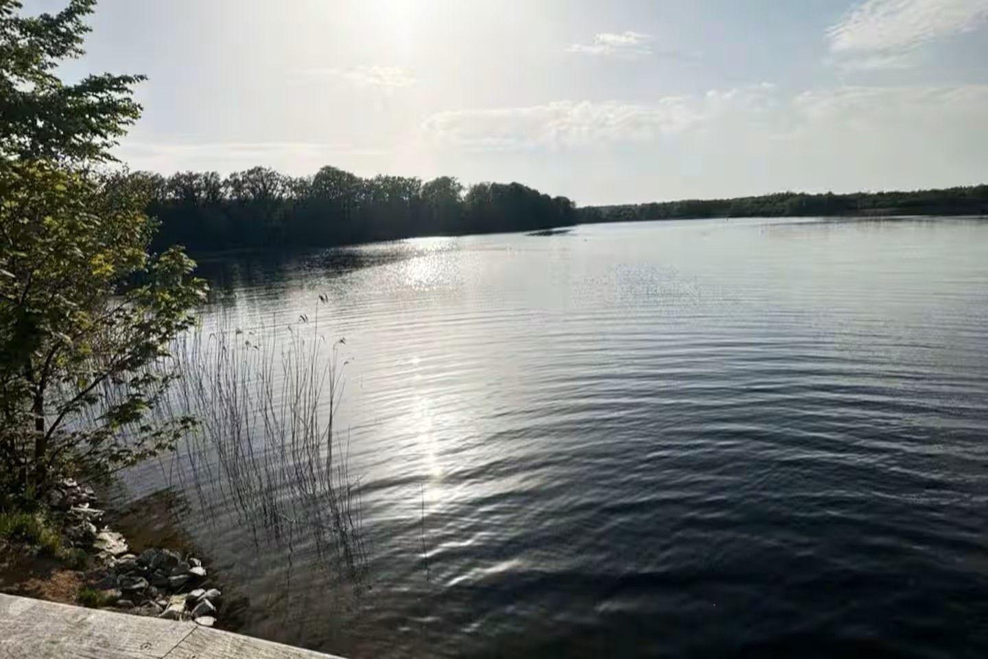 Stiller See mit Uferbereich und Bäumen im Hintergrund. Sonne spiegelt sich auf der Wasseroberfläche.