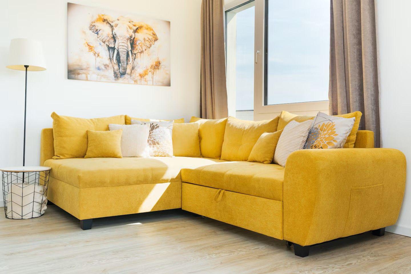 Gelber Ecksofa mit Kissen, Wandbild und Fenster mit Vorhängen.