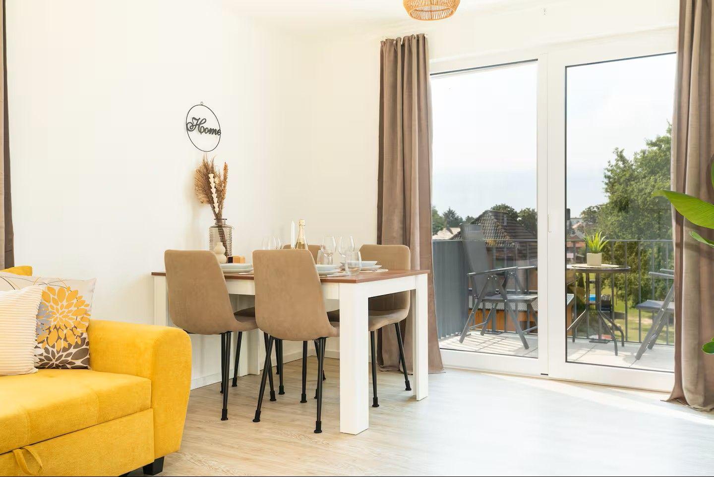 Essbereich mit Tisch und Stühlen, gelber Sofa-Ecke und Balkon mit Sitzgelegenheiten.