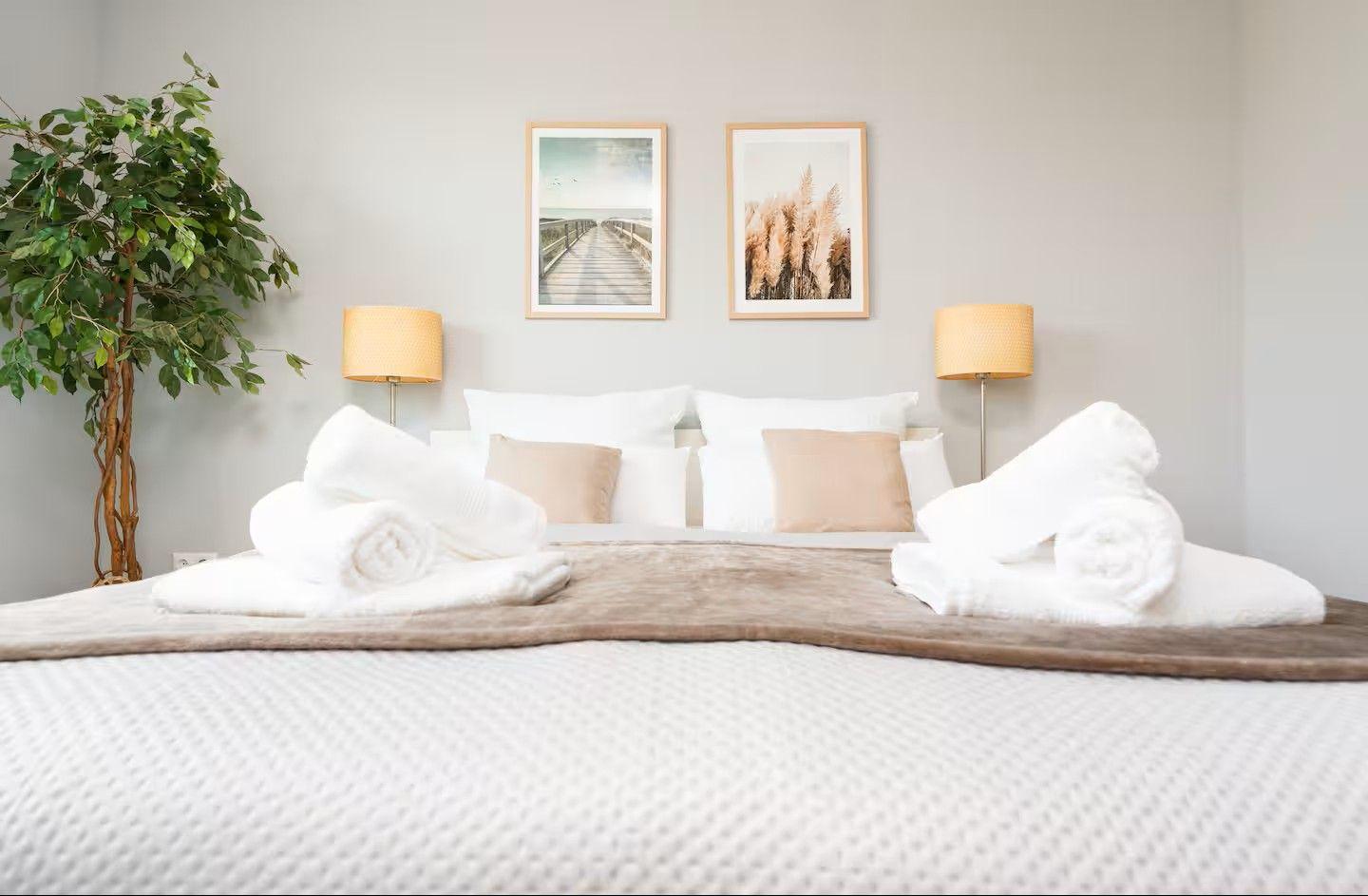 Das Bett mit weißer Bettwäsche, Kissen und aufgerollten Handtüchern. Zwei Bilder und eine Pflanze an der Wand.