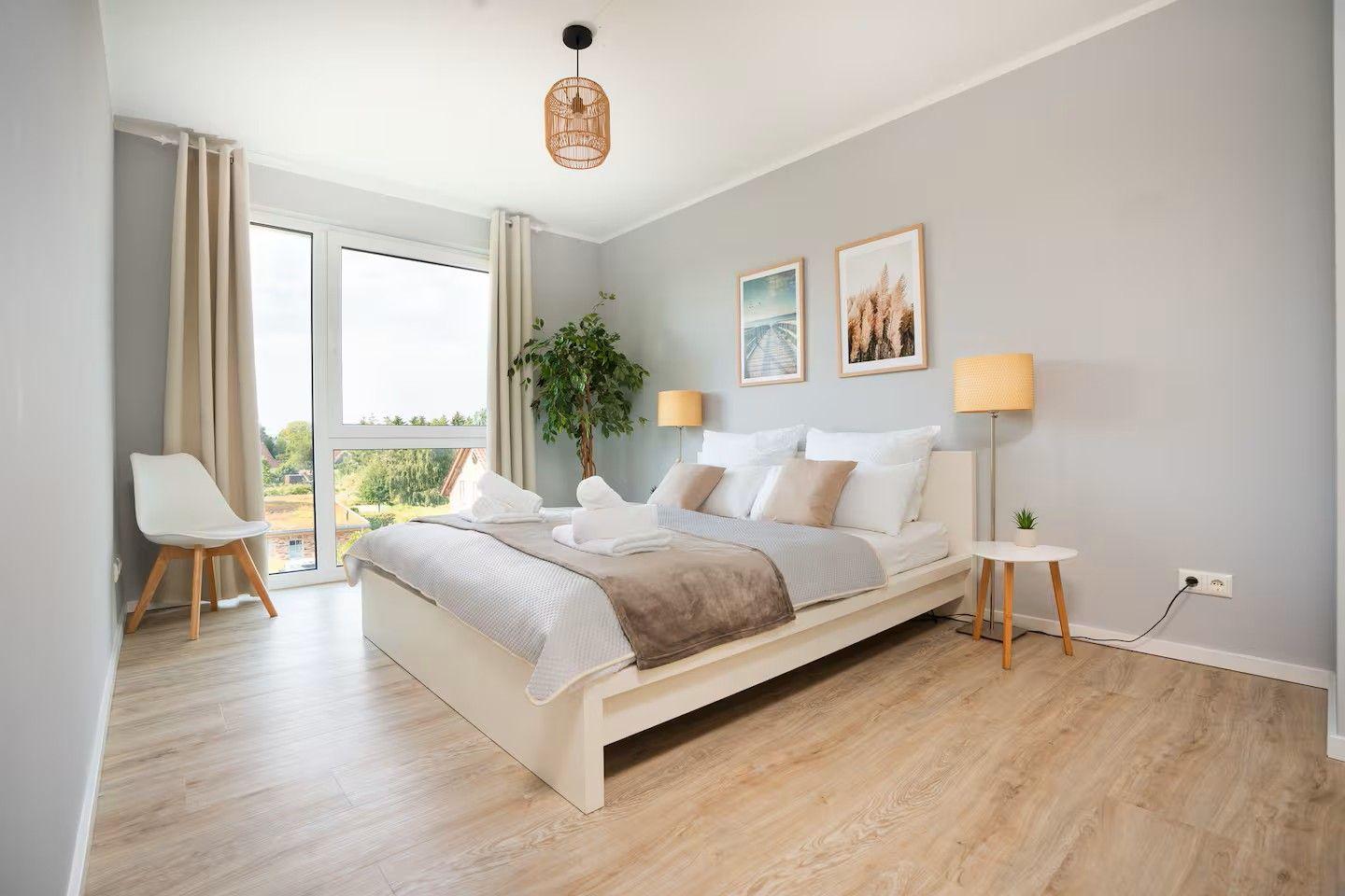 Schlafzimmer mit Bett, Nachttisch, Lampe und großem Fenster. Holzboden und Wandbilder.