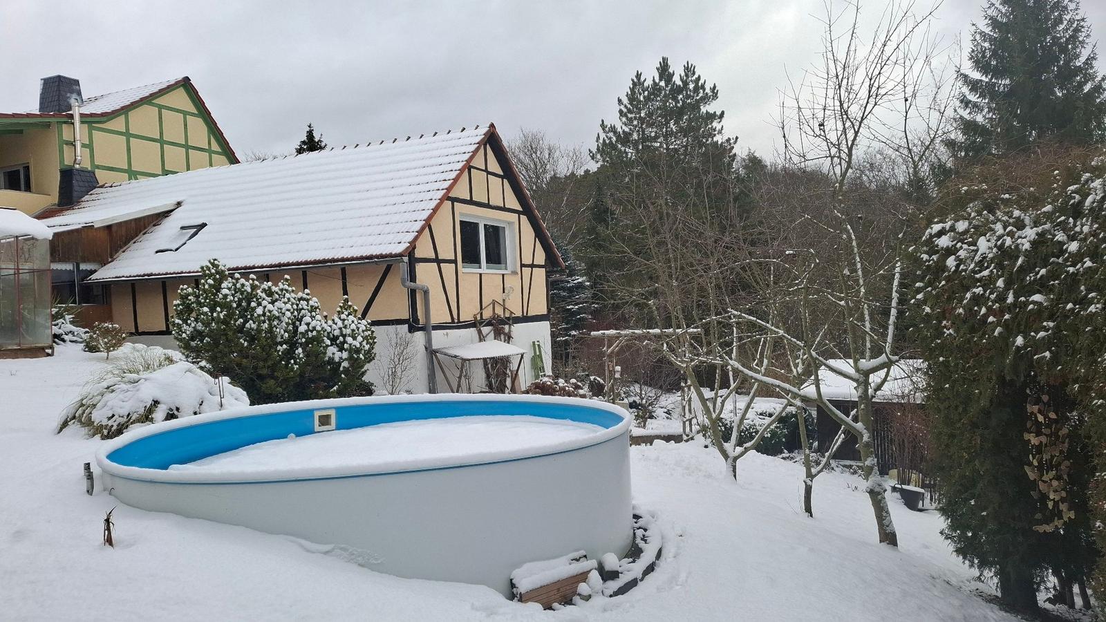 Schneebedeckter Garten mit Pool und Fachwerkhaus im Hintergrund.
