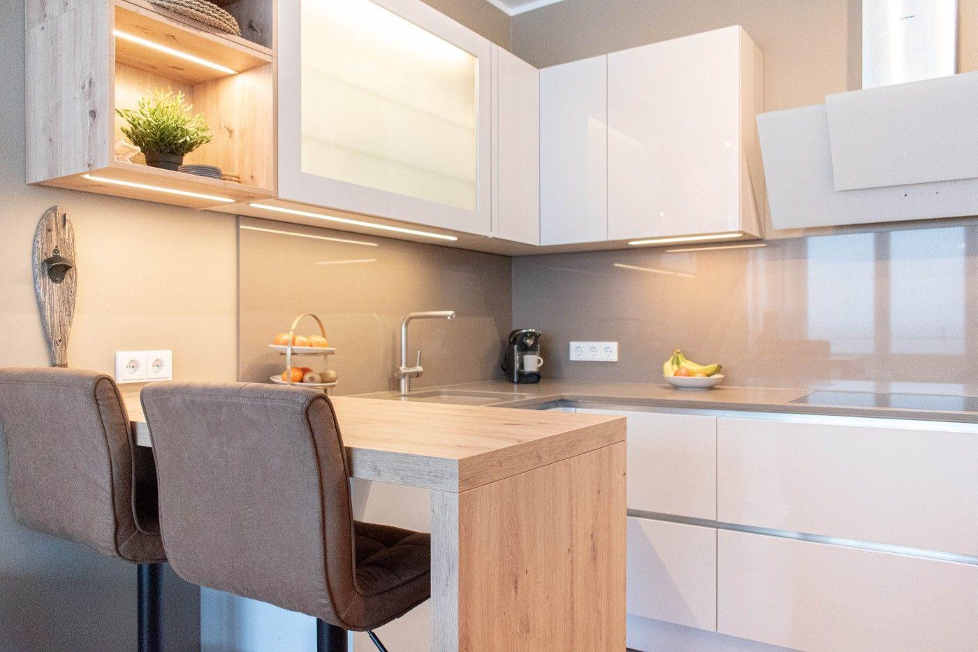 Küche mit Holzinsel, weißen Schränken und Barhockern. Auf der Arbeitsplatte stehen Obst und eine Kaffeemaschine.