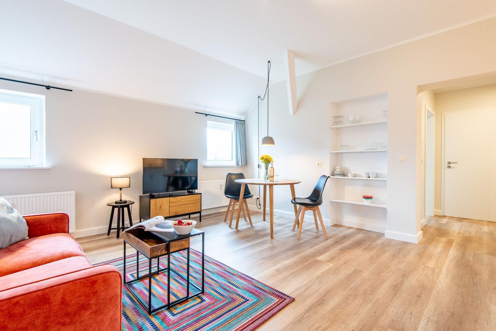 Wohnzimmer mit Sofa, Esstisch und Regal. Holzboden und Fenster sichtbar.