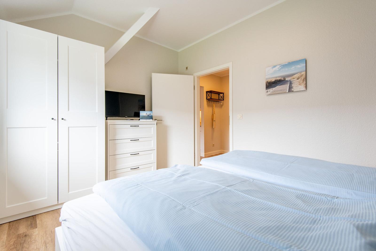 Schlafzimmer mit Doppelbett, Kleiderschrank und Fernseher. An der Wand hängt ein Strandbild.