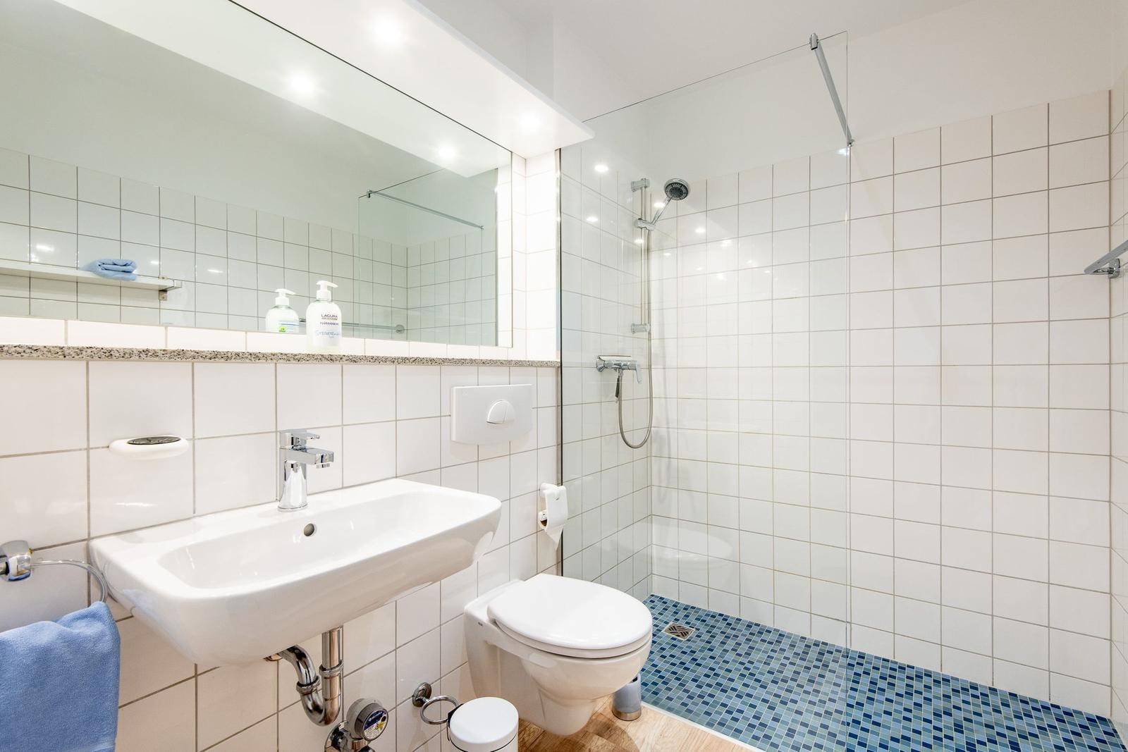 Badezimmer mit Dusche, Waschbecken und Toilette. Fliesenboden und Spiegel.