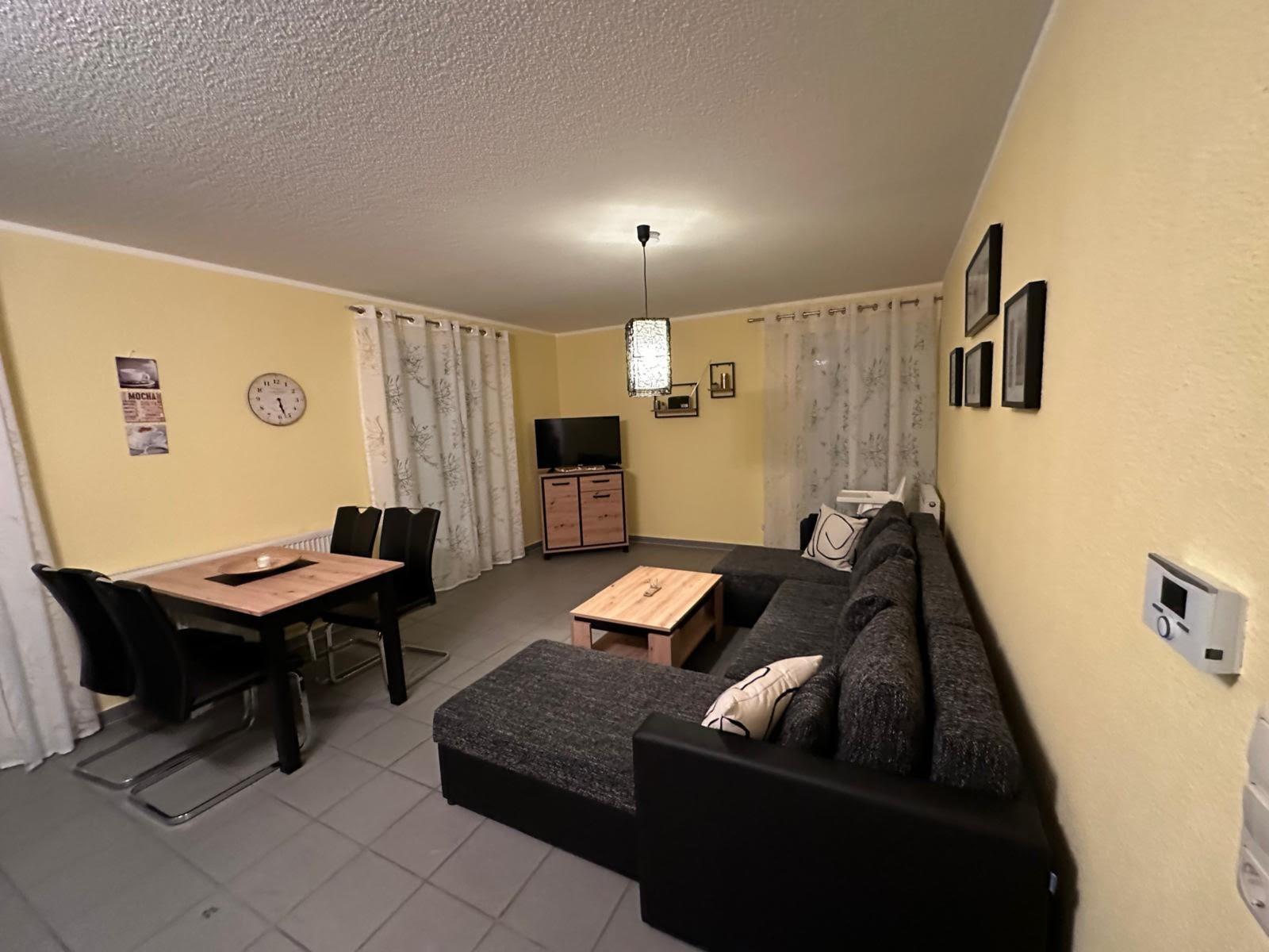 Wohn- und Essbereich mit Sofa, Tisch, Fernseher und Vorhängen.