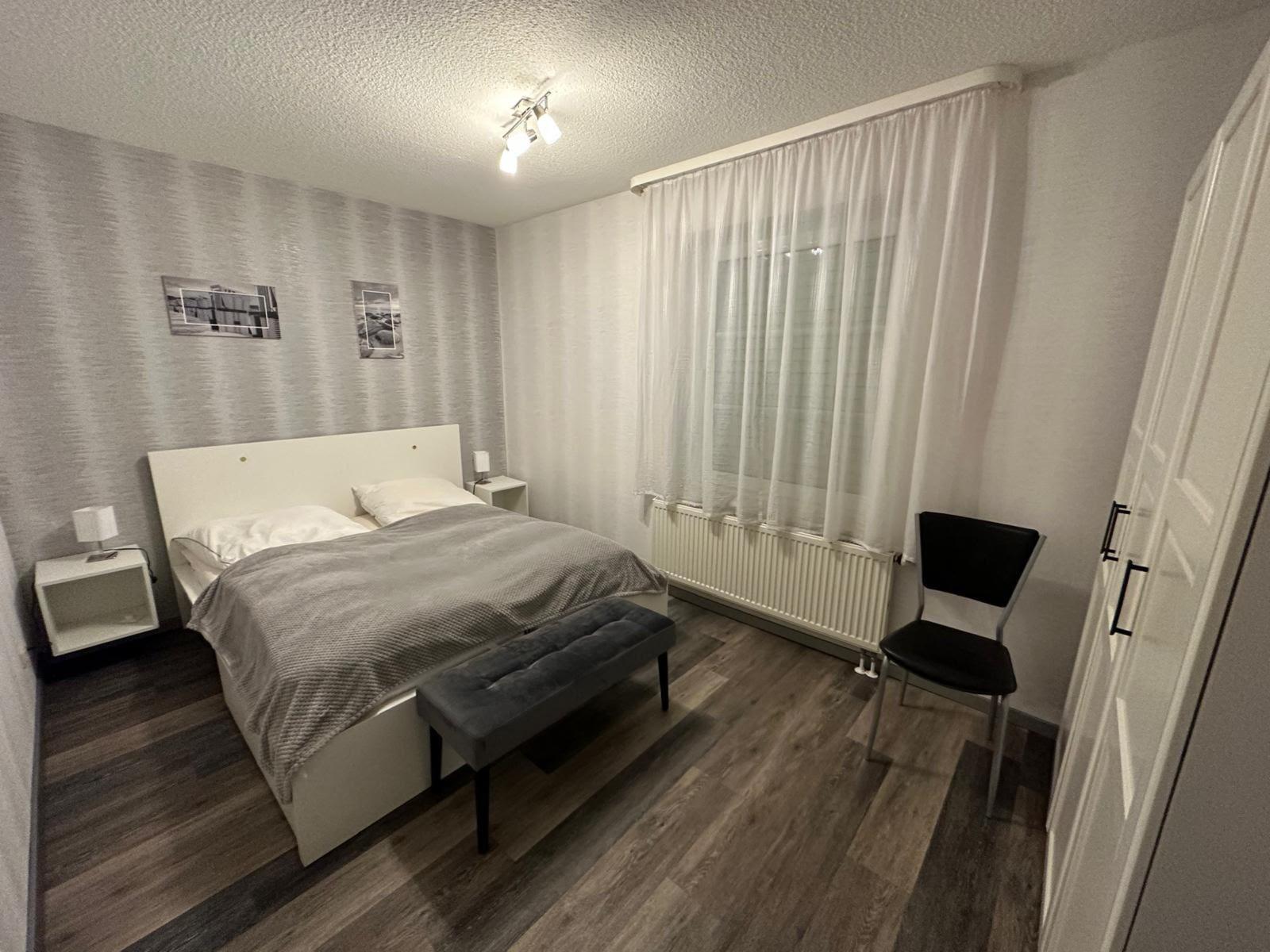 Schlafzimmer mit Bett, Nachttischen, Stuhl und Fenster mit Vorhängen.