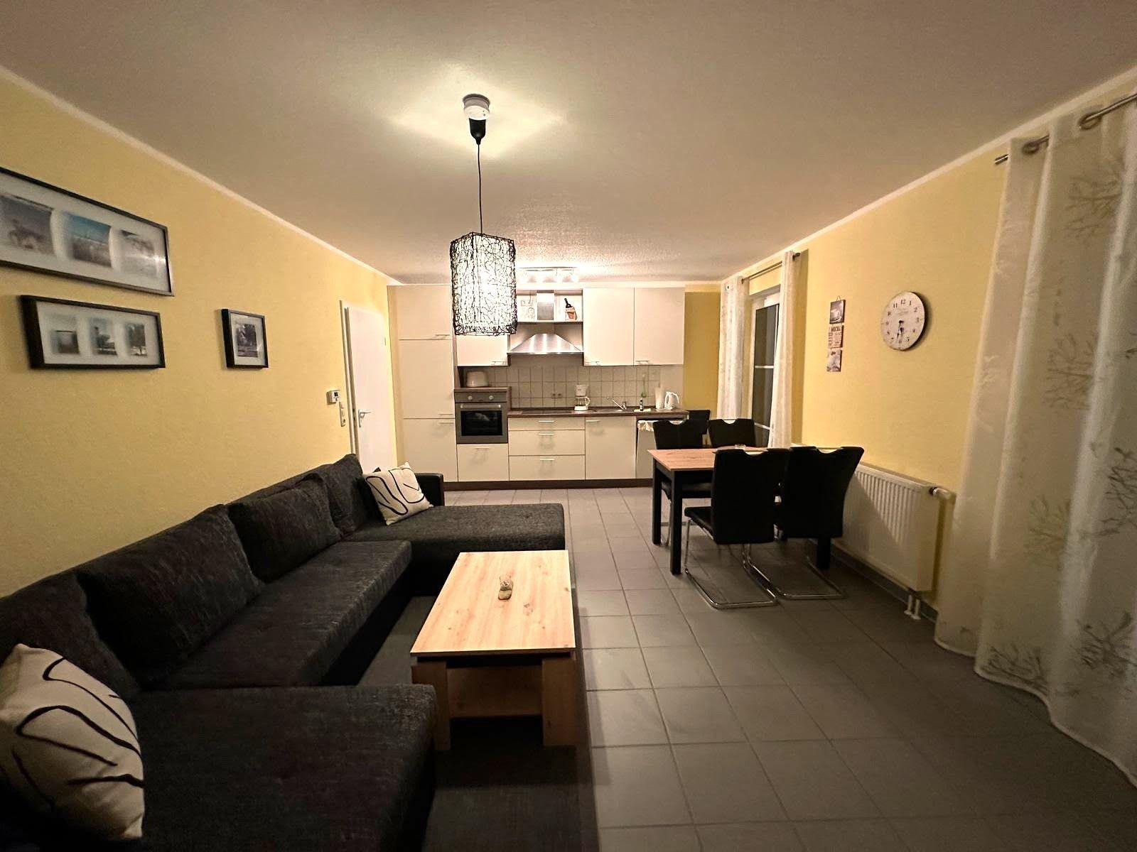 Wohn- und Essbereich mit Küche, Sofa, Tisch und Stühlen. Wand mit Bildern und Uhr.
