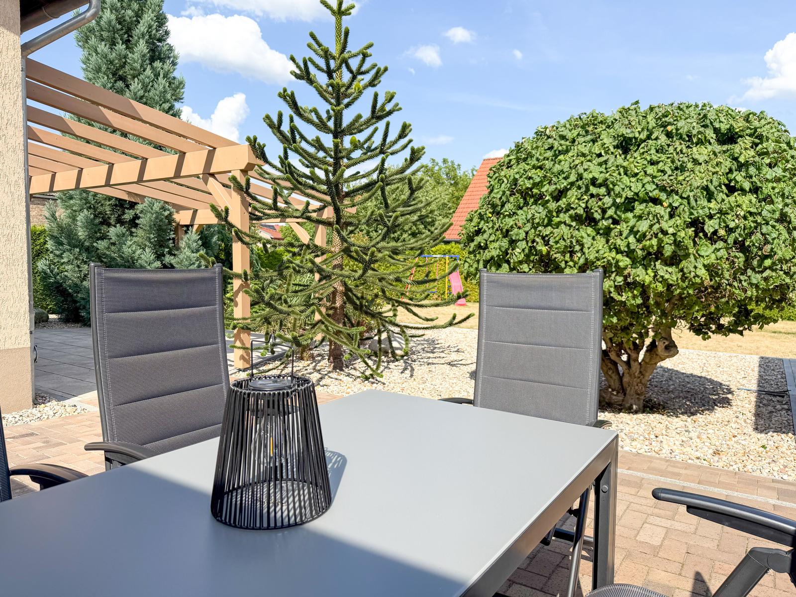Terrasse mit Tisch und Stühlen, umgeben von Bäumen und Sträuchern unter blauem Himmel.