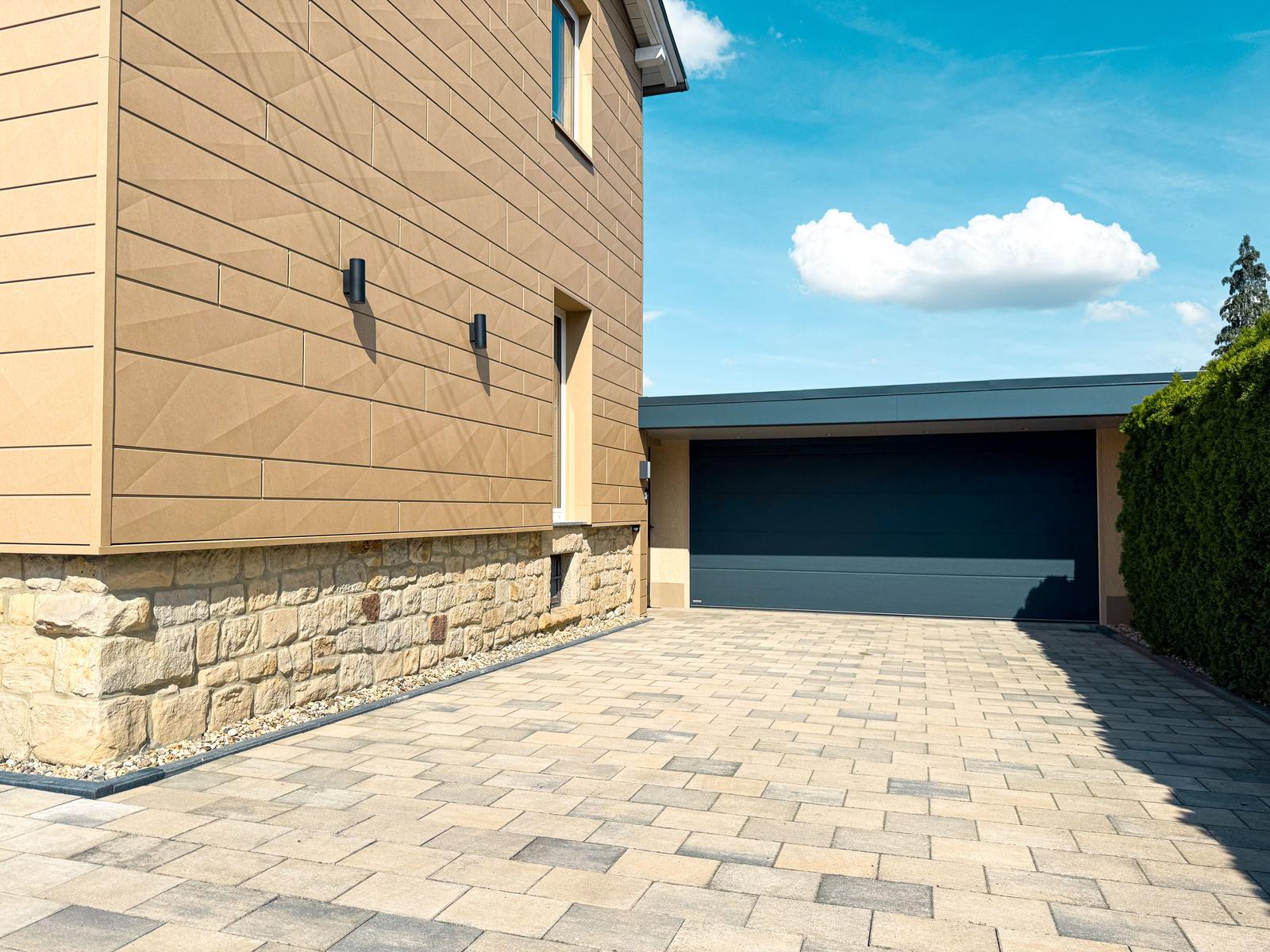 Moderne Villa mit Garagenbereich und gepflastertem Hof unter blauem Himmel.