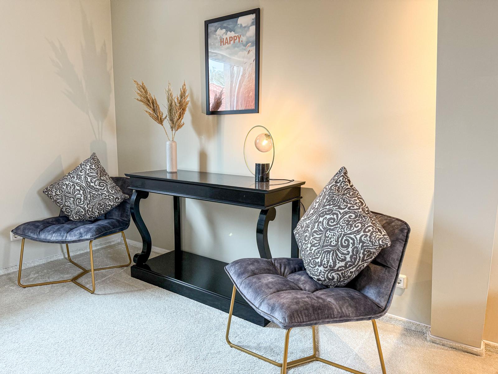 Zwei Sessel mit Kissen stehen um einen schwarzen Tisch herum. An der Wand hängt ein Bild.