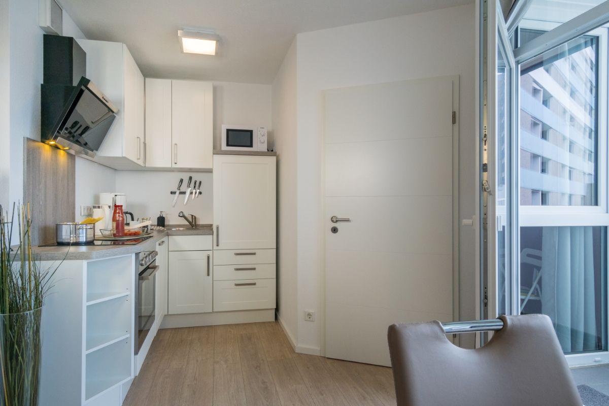 Küche mit weißen Schränken, Herd und Dunstabzug. Tür und Balkon sind sichtbar.