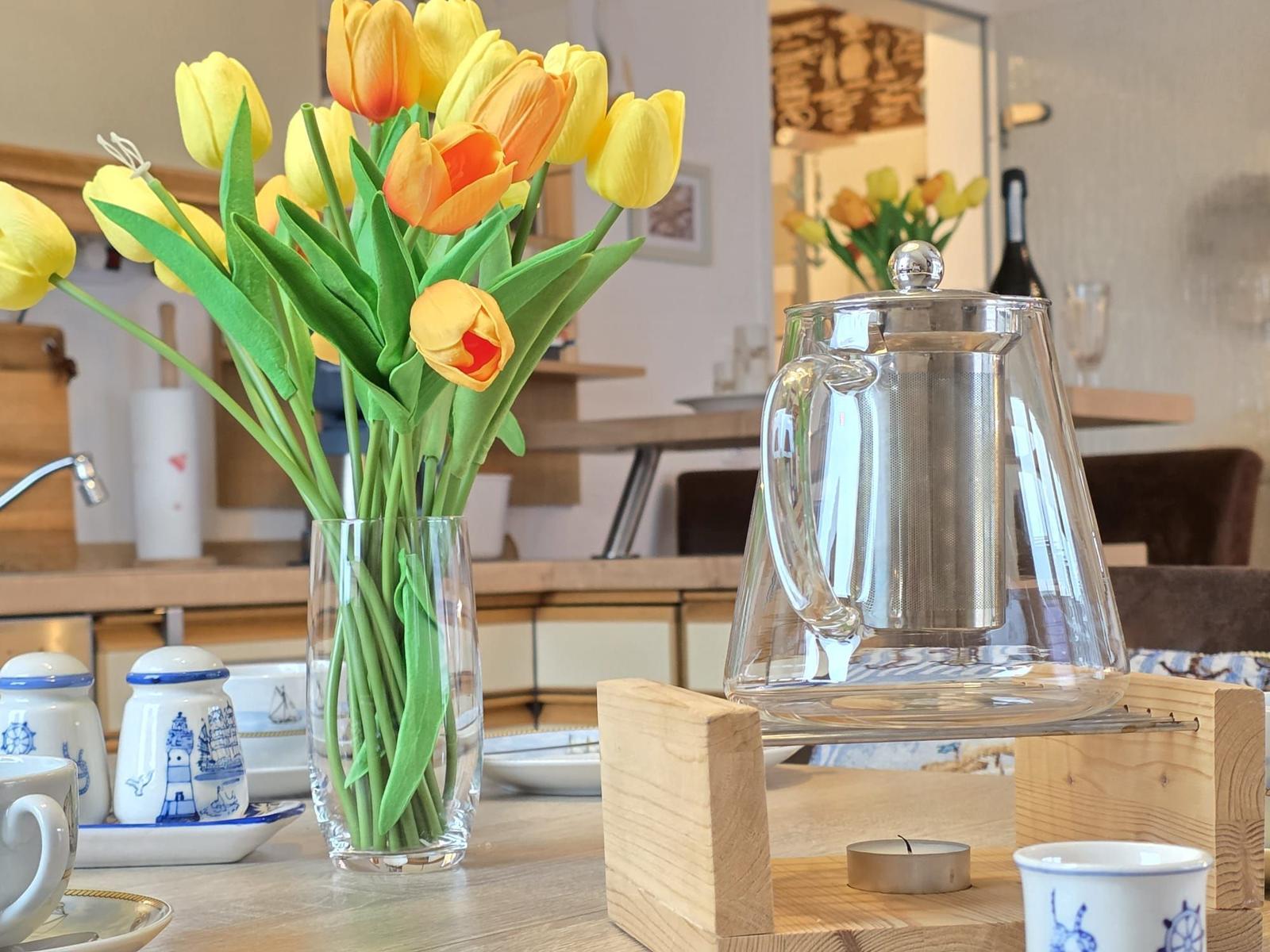 On the table, there is a vase with tulips and a glass kettle on a wooden stand.