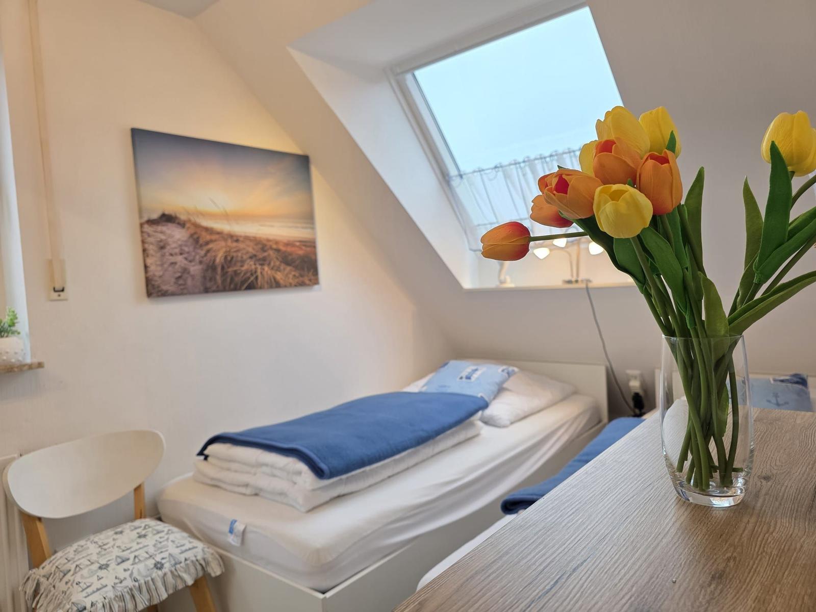 A bedroom with sloped ceilings, bed, chair, and tulips in vase.