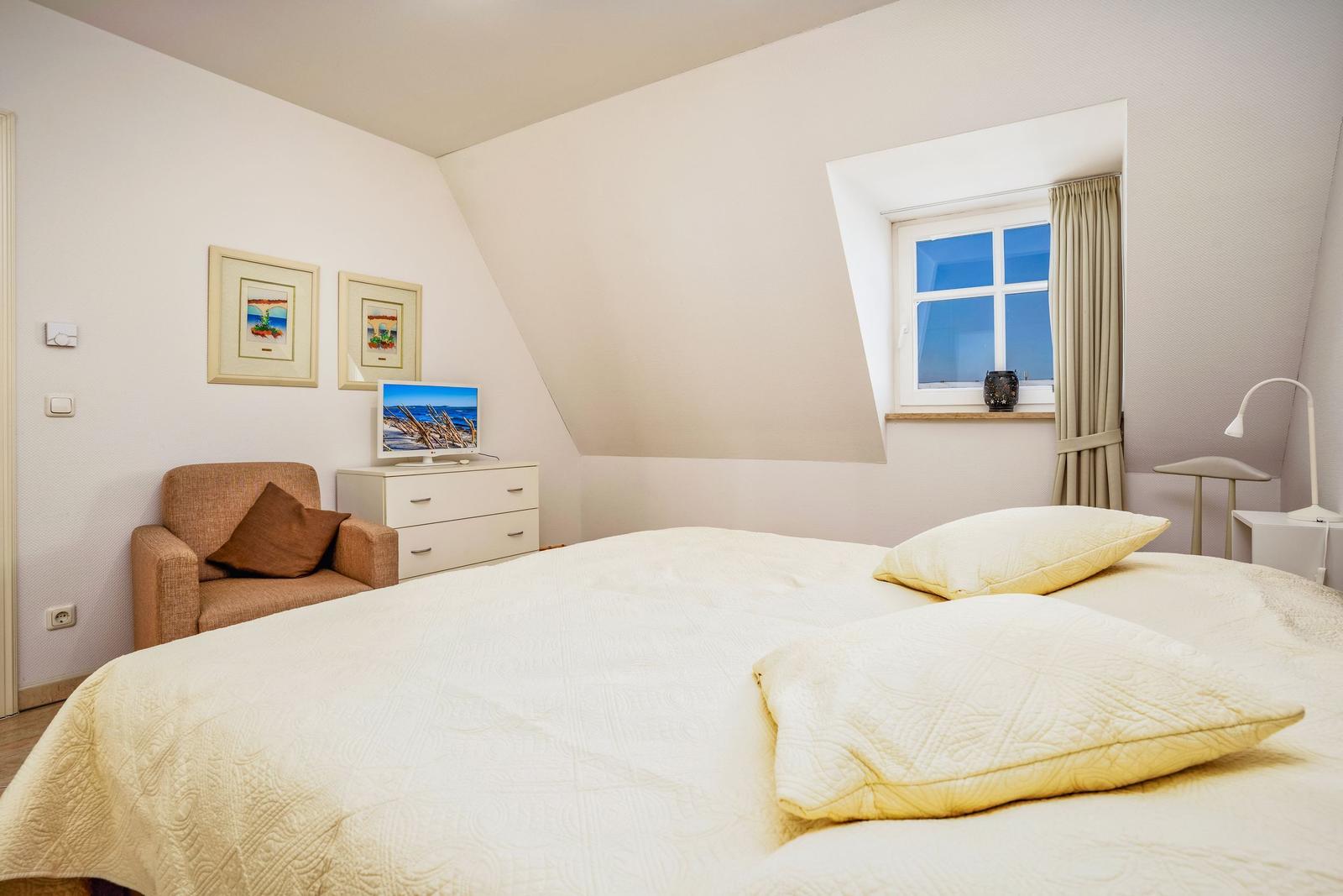 Schlafzimmer mit Bett, Sessel, Schrank und Fenster. Wandbild und Nachttisch sichtbar.