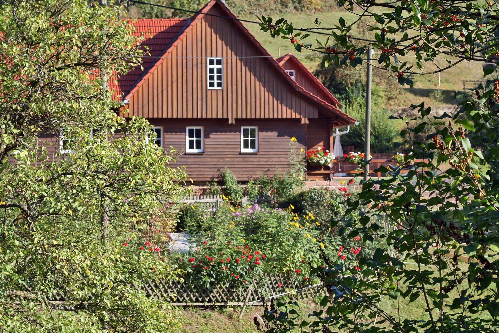 Holzhäuschen mit rotem Dach, umgeben von Blumen und Bäumen.