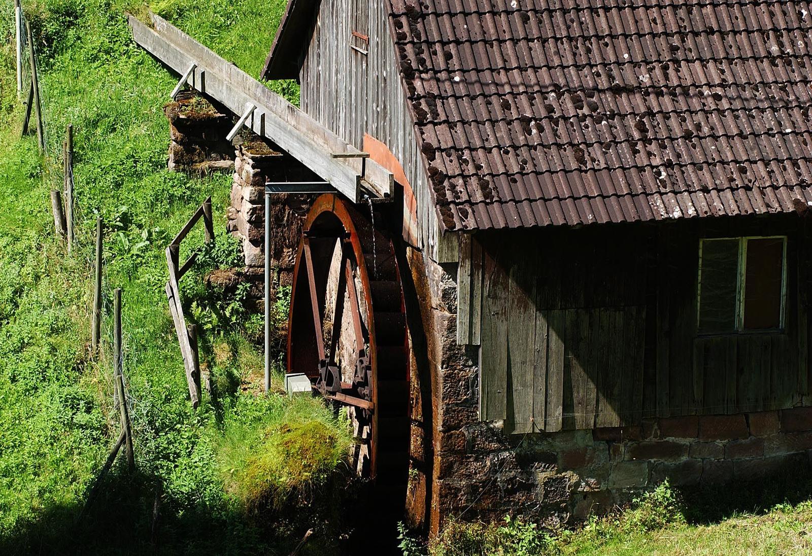 Alte Wassermühle mit Holzrad und Steinmauer an grüner Hanglage.
