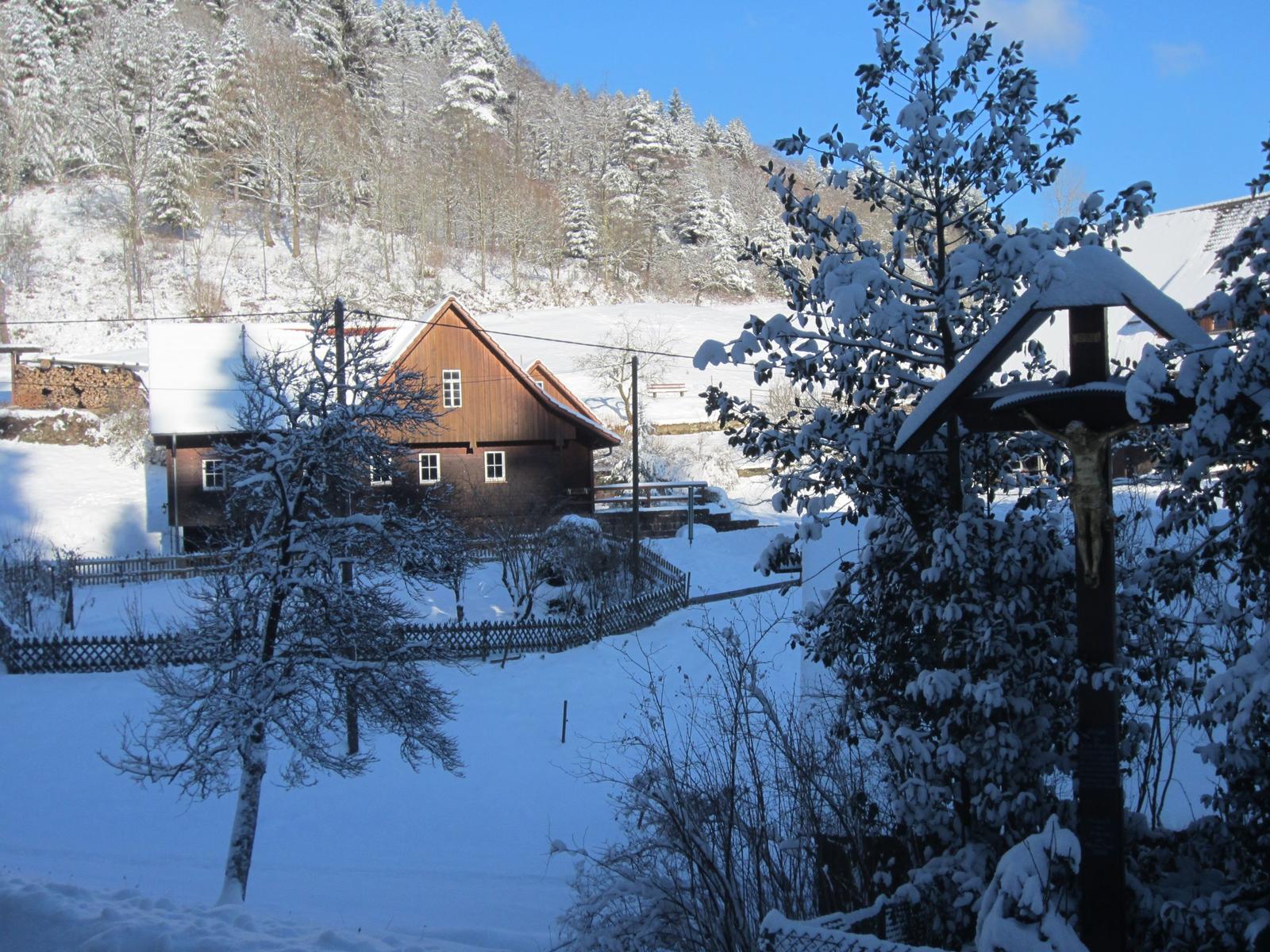 Schneebedecktes Haus mit Holzveranda und umliegenden Bäumen im Winter.