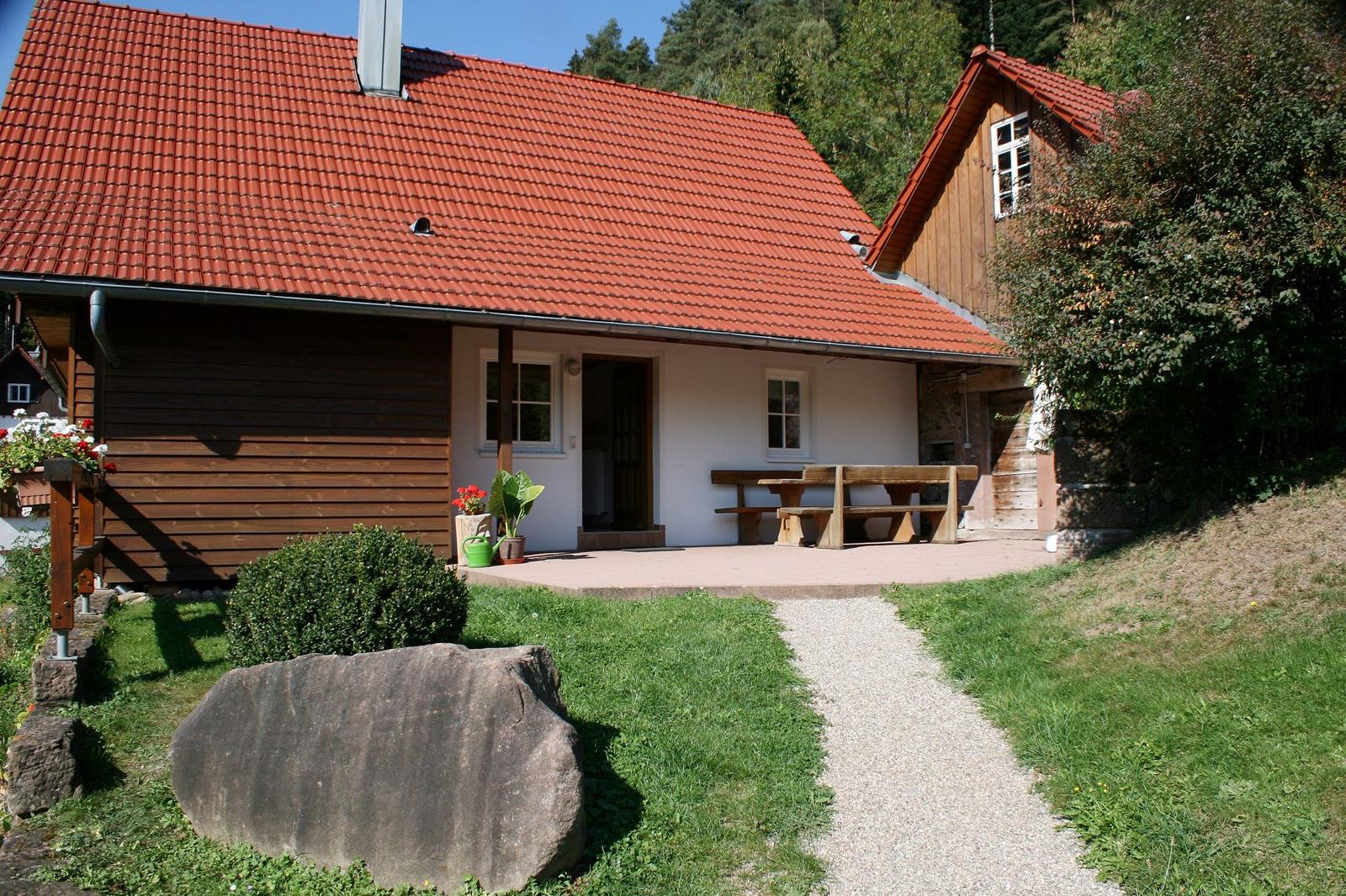 Haus mit rotem Dach, Terrasse und Holztisch. Gras, Steinweg und Bäume umgeben es.