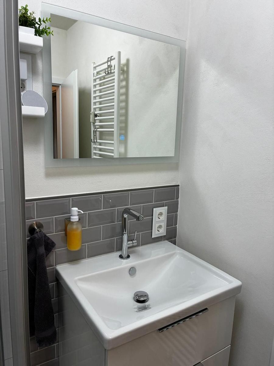 Waschbecken mit Spiegel, Handtuchhalter und Seifenspender an grauer Fliesenspiegelwand.