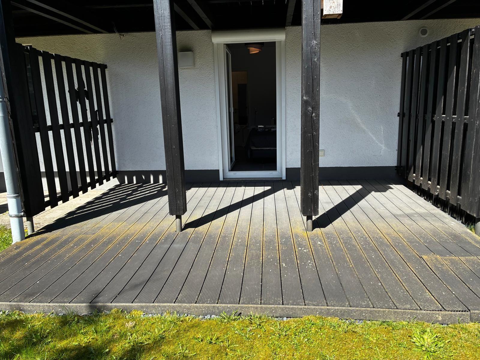 Terrasse mit Holzdielen und schwarzem Zaun, Tür führt in das Haus.