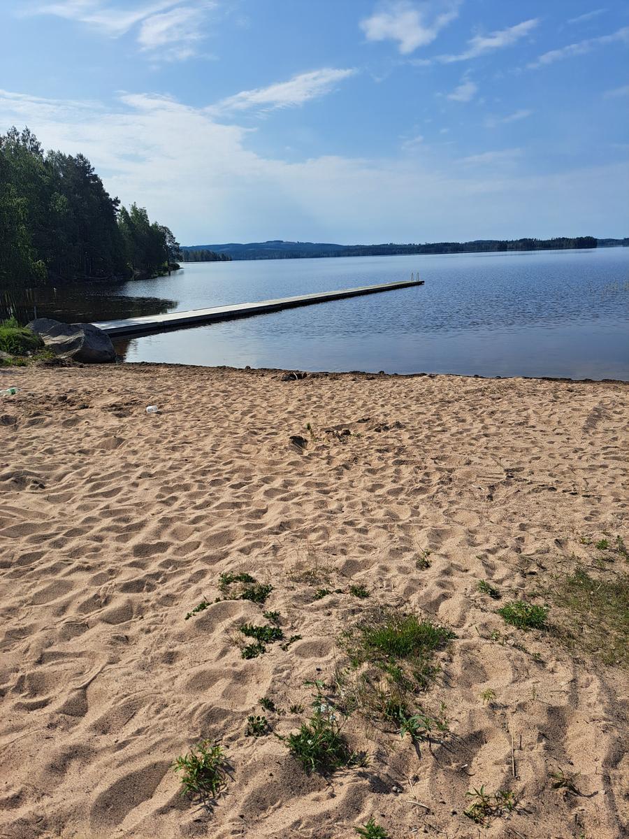 Sandstrand mit Holzsteg zum See. Wald im Hintergrund unter blauem Himmel.