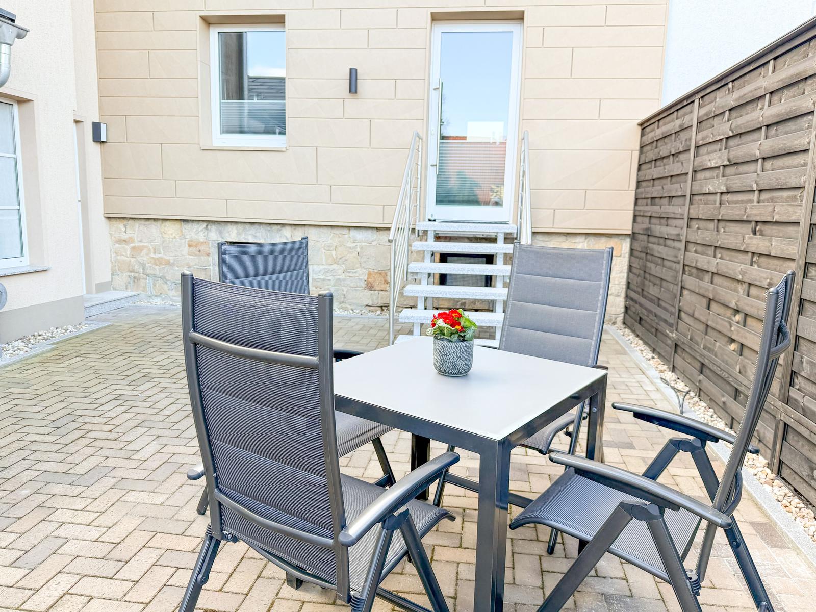 Terrasse mit Tisch und vier Stühlen, Pflanze darauf, Holzzaun und Hauswand im Hintergrund.