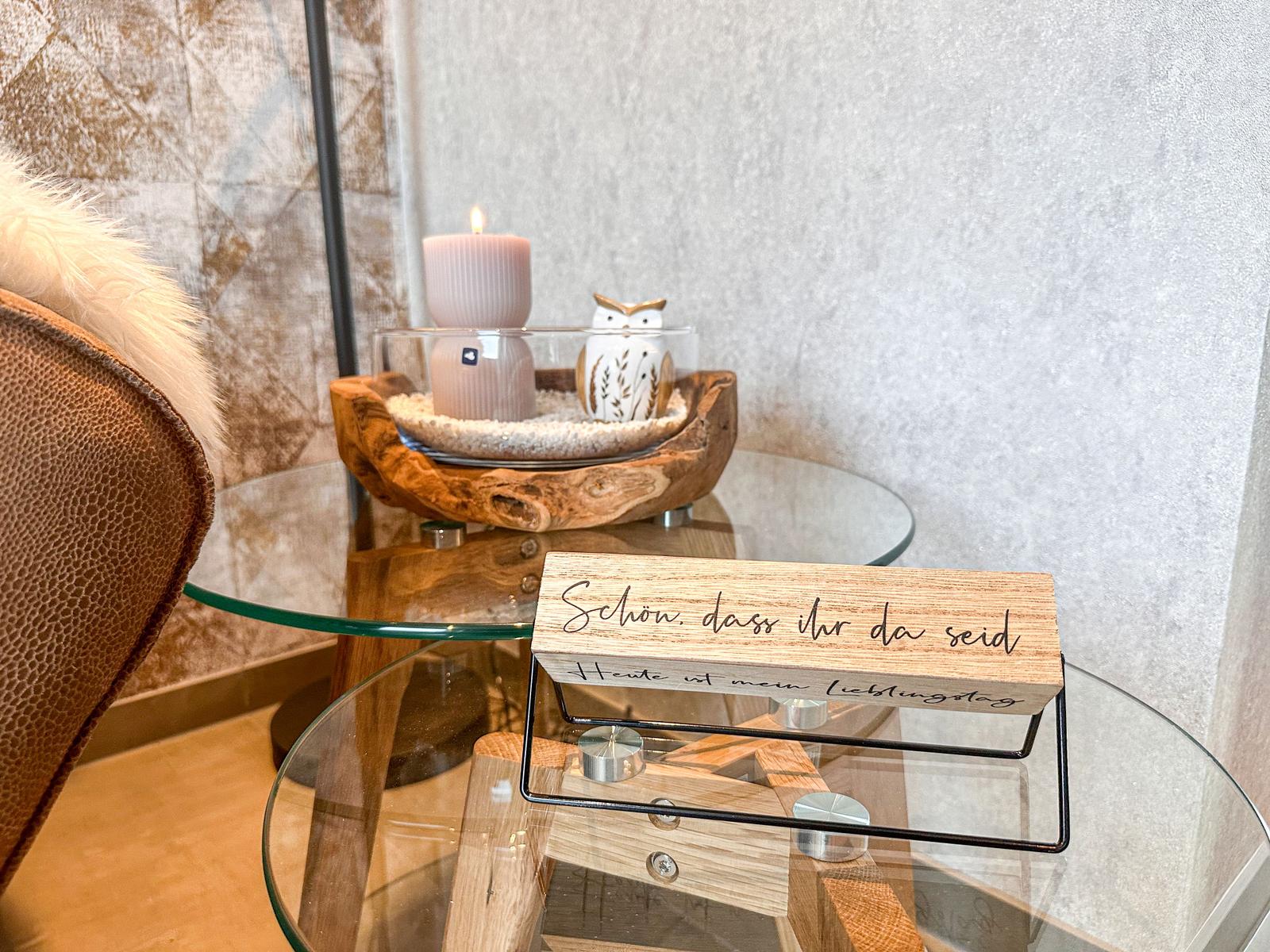 Auf einem Glas-Tisch steht eine Kerze, eine Eule und ein Holzschild mit Schrift.