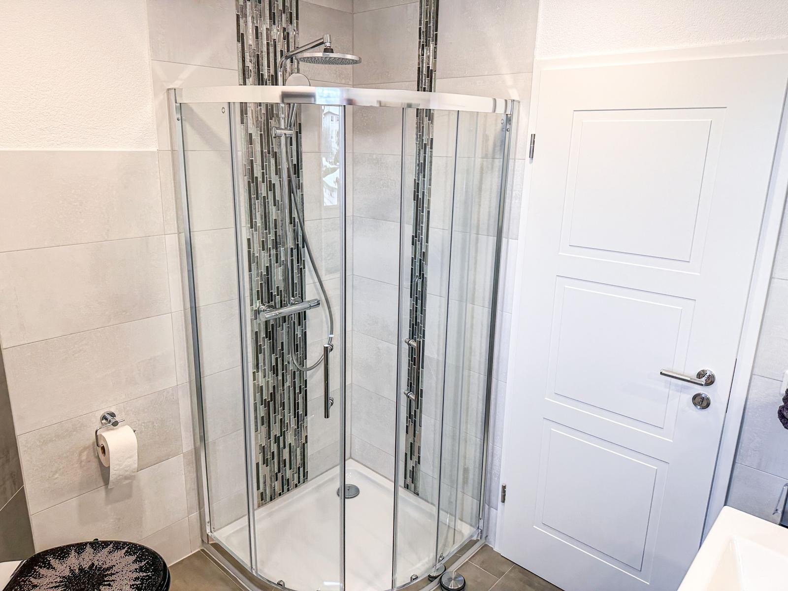 Duschkabine mit Glaswand, Mosaikfliesen und Duschkopf. Weiße Tür und Toilettenpapierhalter.