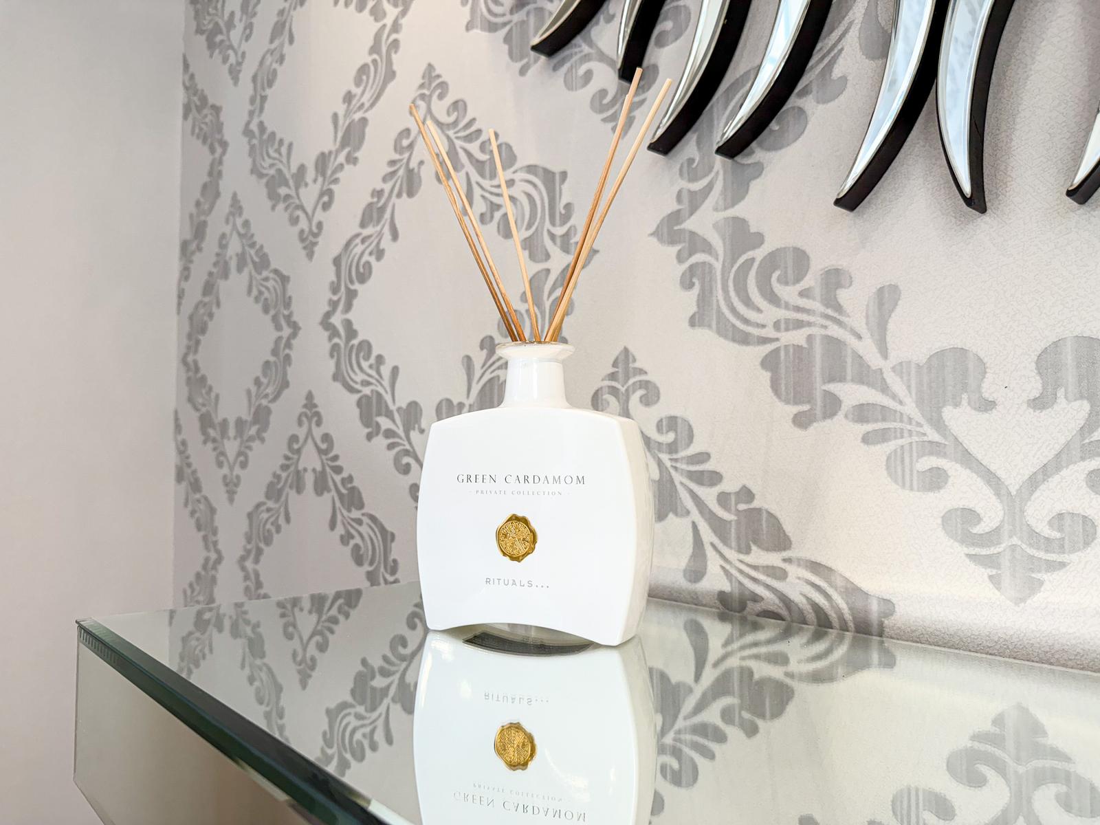 Weißer Duftdiffusor mit Holzstäbchen auf glänzender Oberfläche vor gemustertem Wandbehang.