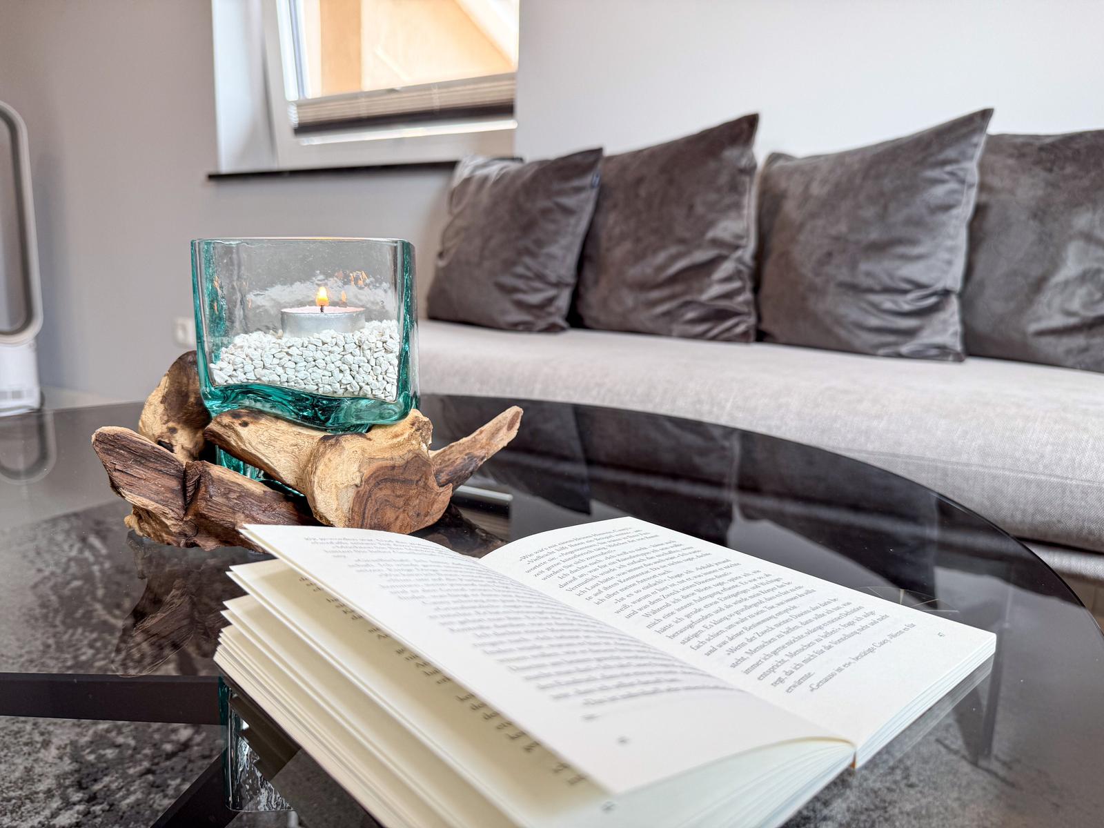 Ein Glas-Tisch mit Buch und Kerze auf Holzdekoration. Hintergrund: Sofa mit Kissen.