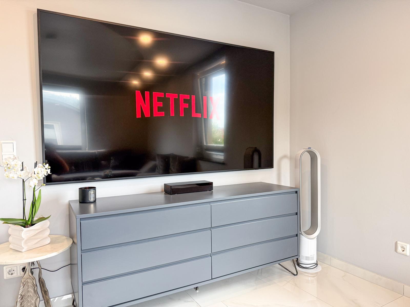 Großer Fernseher mit Netflix-Symbol, graue Kommode, weißer Staubsauger und Orchidee auf Tisch.