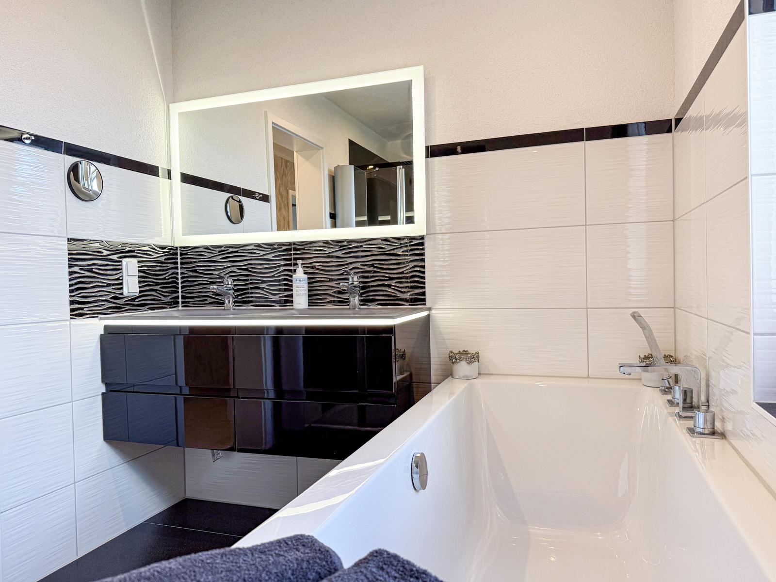 Badezimmer mit Badewanne, Waschbecken und Spiegel. Wandfliesen in Weiß und Schwarz.