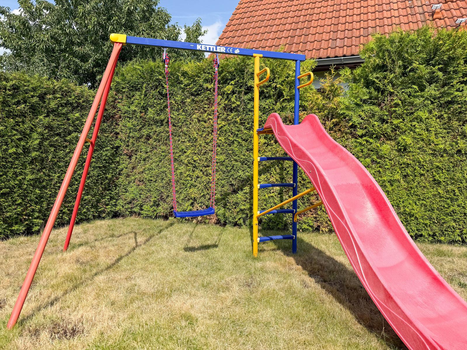 Klettergerüst mit Rutsche und Schaukel im Garten. Hintergrund: grüne Hecke und Hausdach.