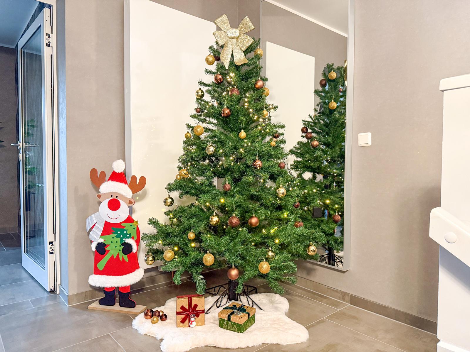 Weihnachtsbaum mit goldenen Kugeln und Schleife, daneben Rentierfigur und Geschenke auf Fellteppich.