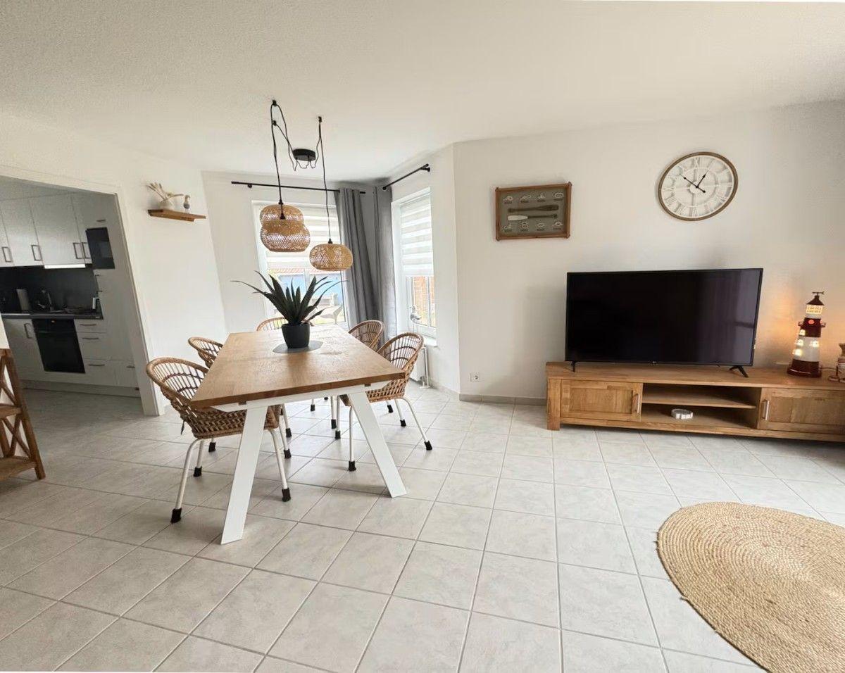Living and dining area with wooden table, TV, and kitchen area in the background.