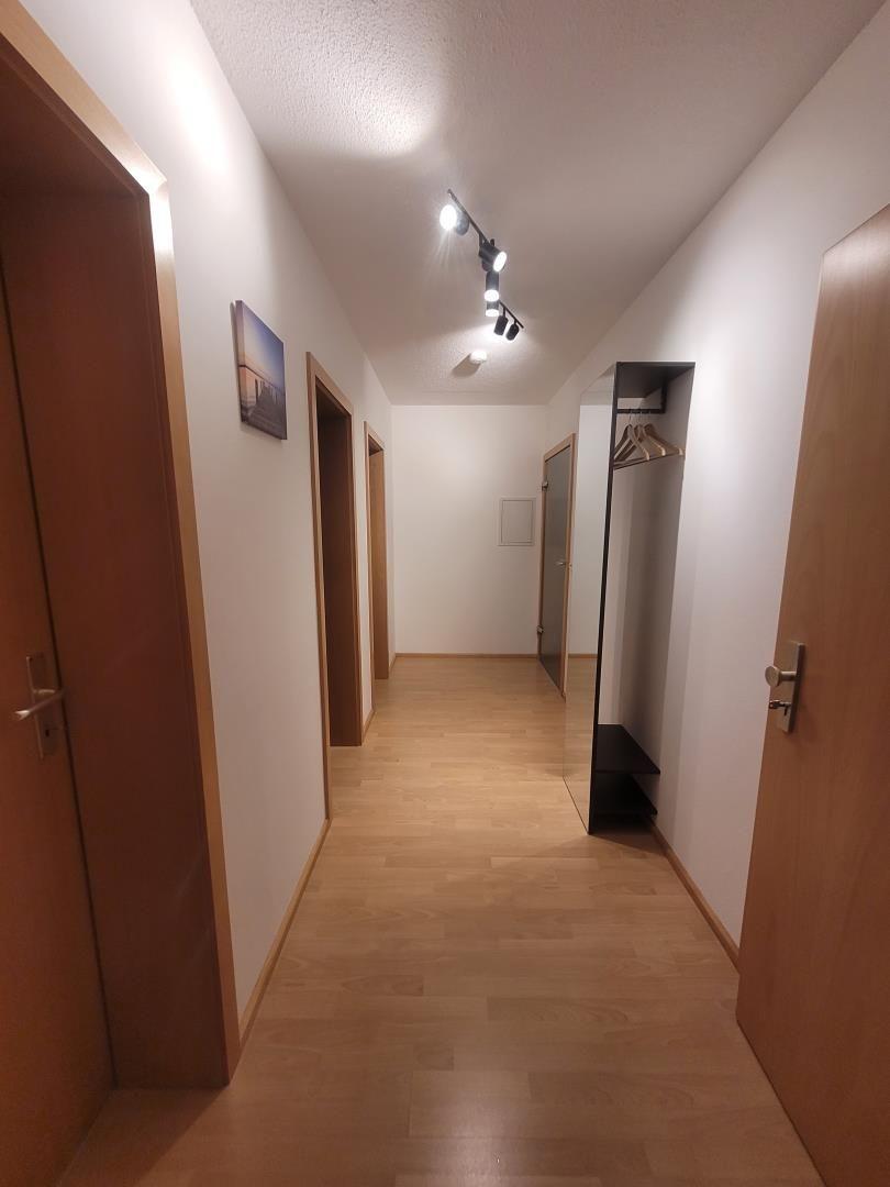 Hallway with wooden floor, doors, and coat rack. Ceiling lights and wall picture visible.