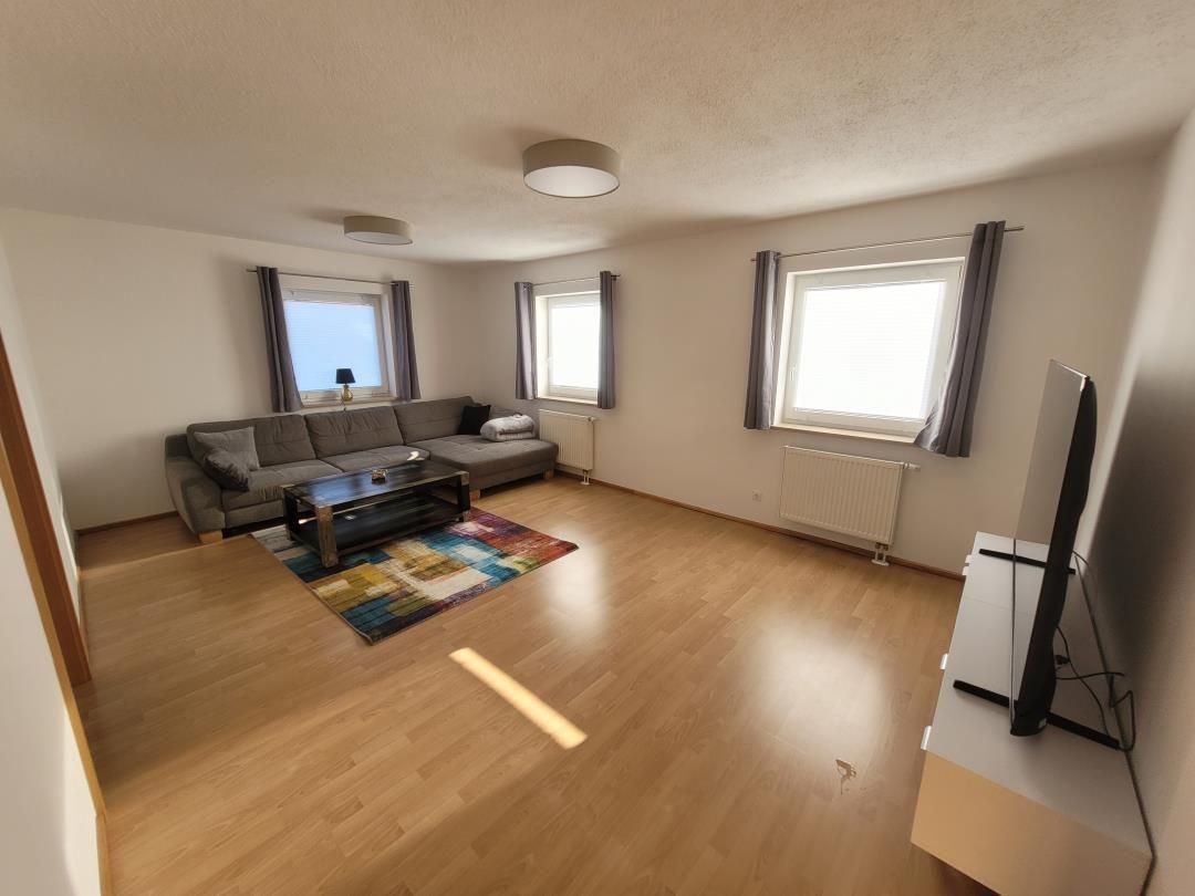 Living room with sofa, coffee table, and TV. Wooden floor and windows with curtains.