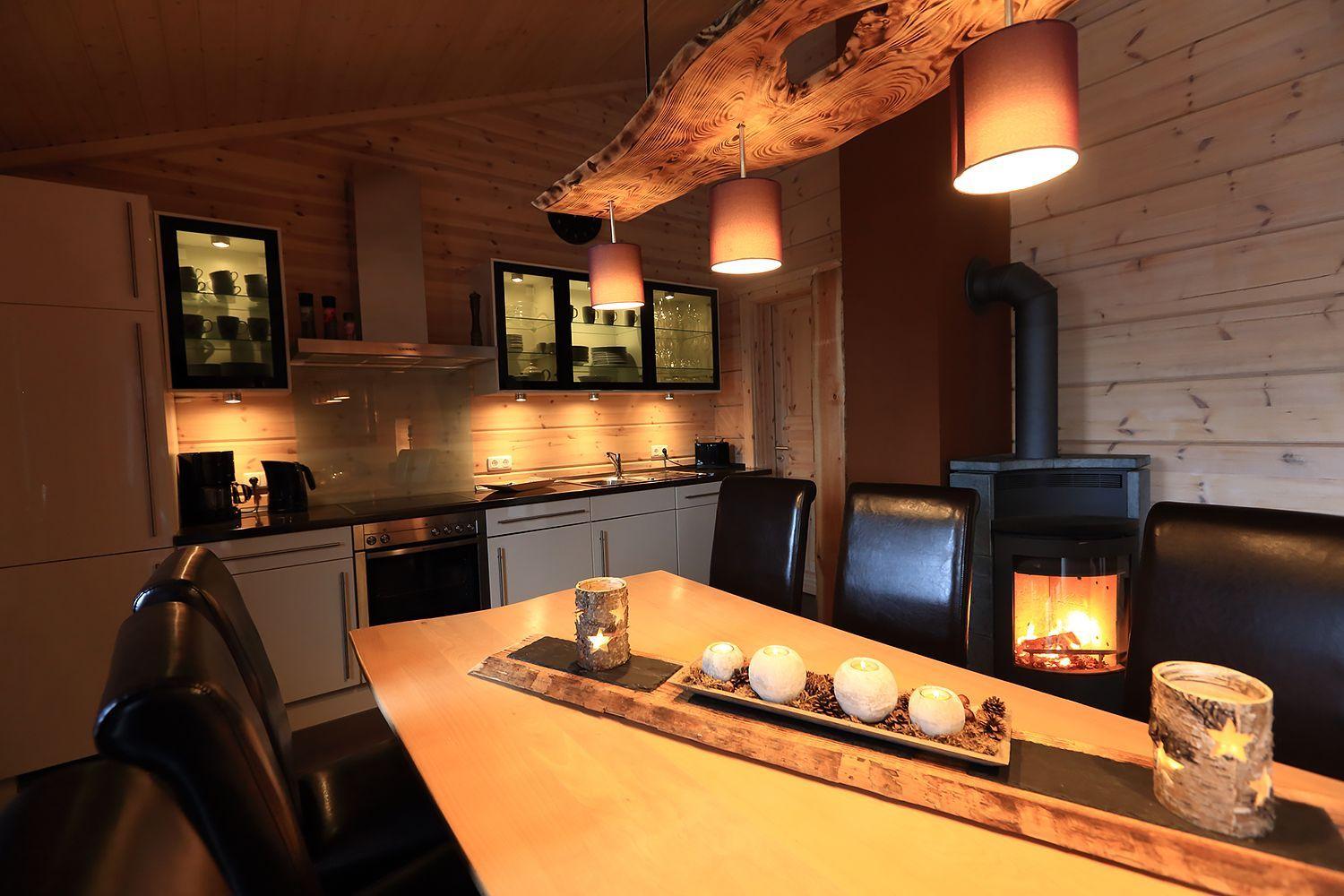 Essbereich mit Holztisch, schwarzen Stühlen und Kamin. Küche mit Holzwand und Glasvitrinen.