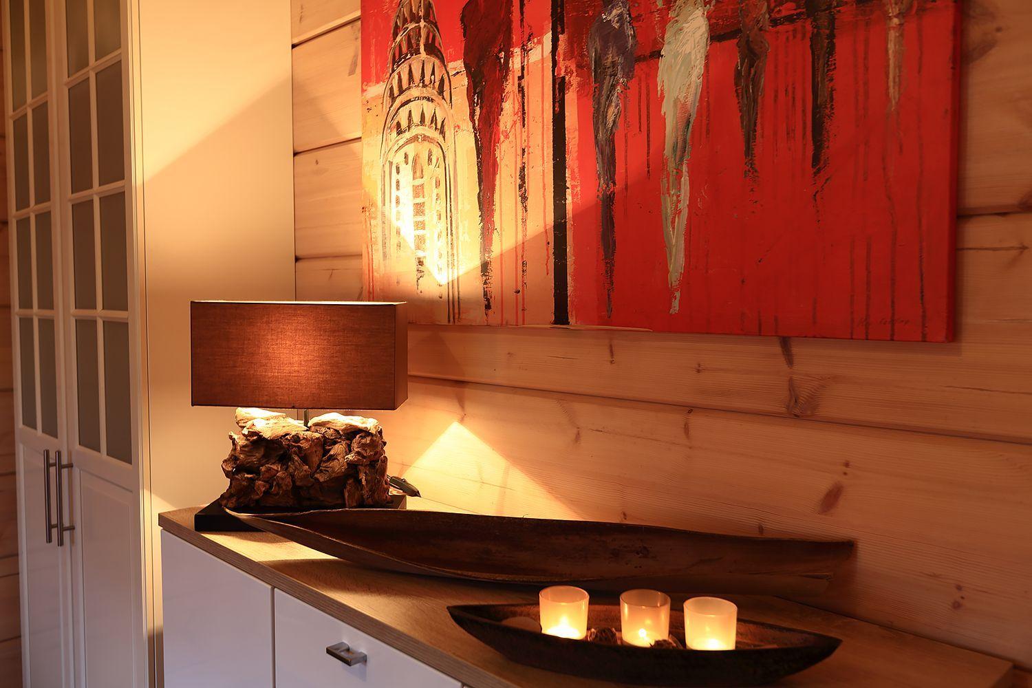 Holzwand mit abstraktem Gemälde, Lampe auf Sideboard und Kerzen in Holzschale.