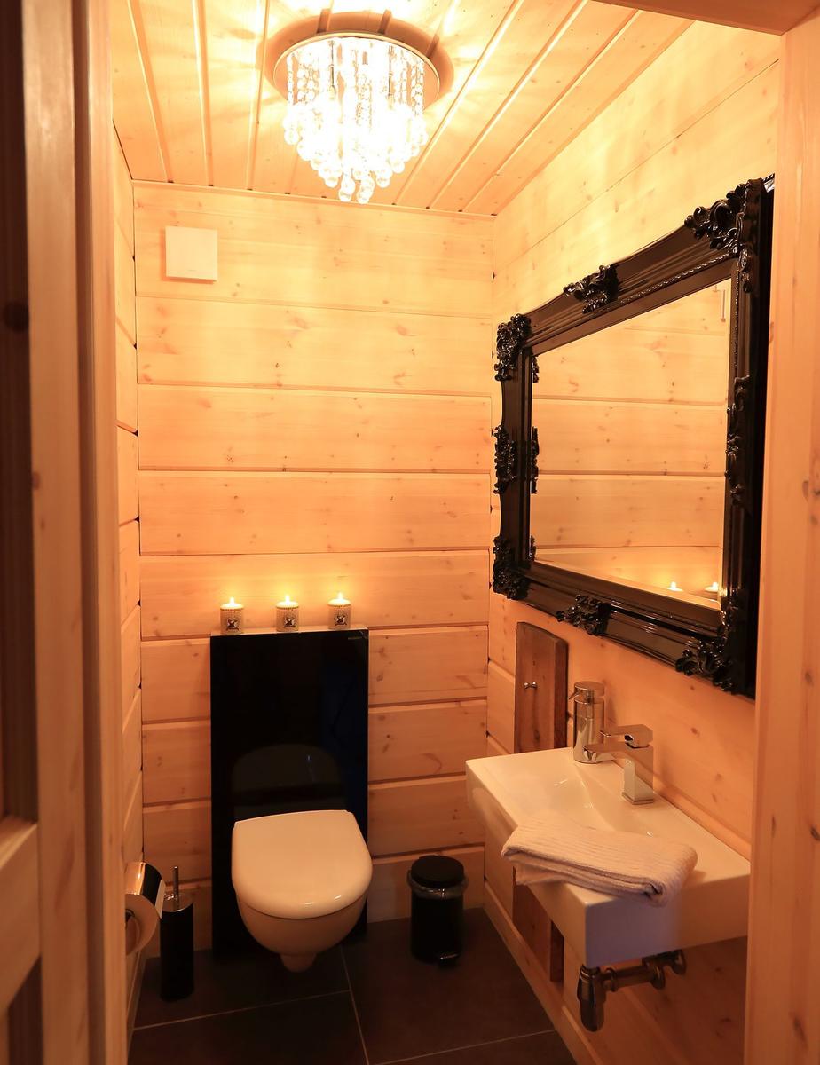Holzbadezimmer mit Toilette, Waschbecken und Spiegel. Deckenleuchte und Kerzen sorgen für Licht.