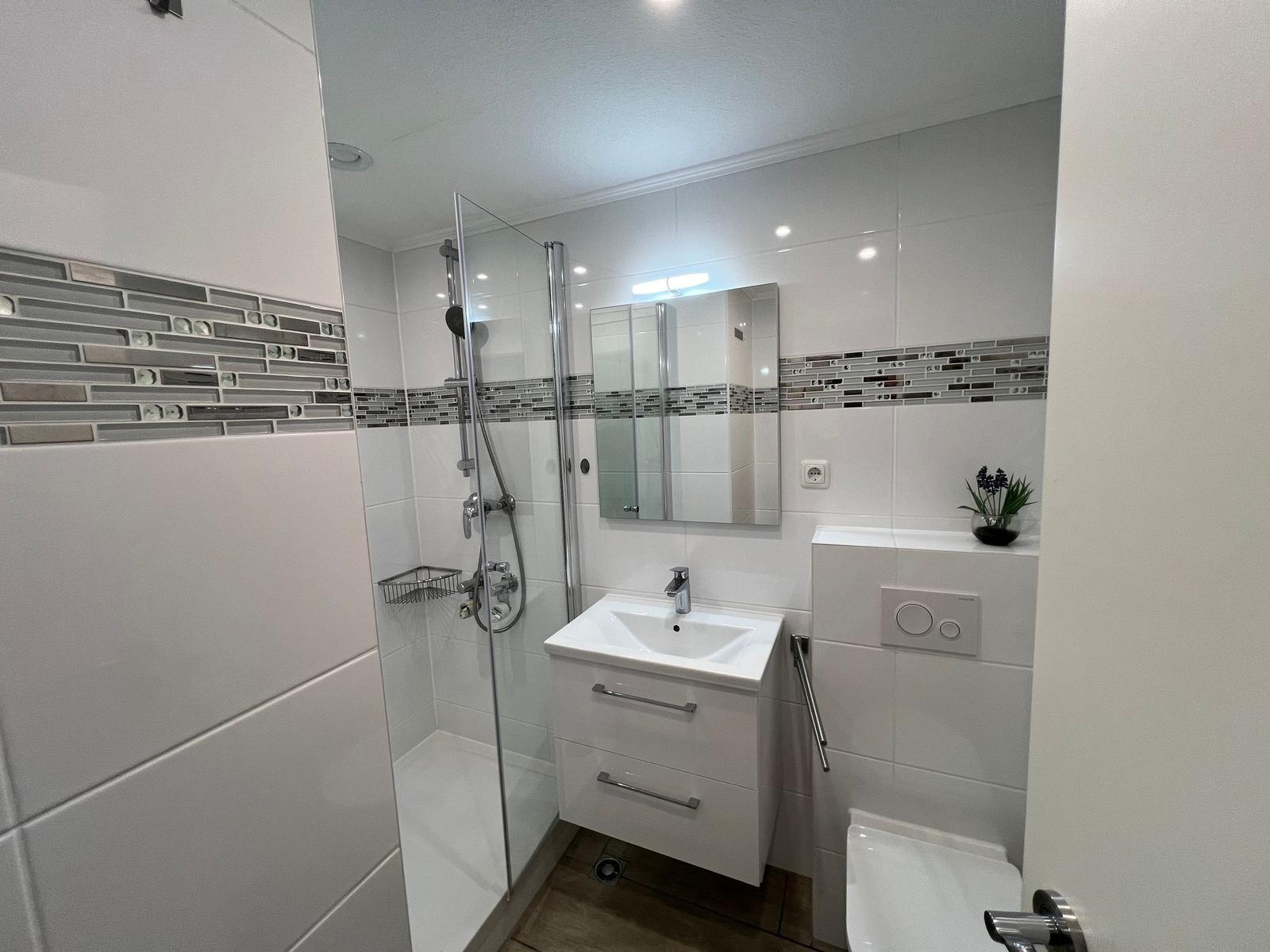 Badezimmer mit Dusche, Waschbecken und WC. Weiße Fliesen und Glaswand.