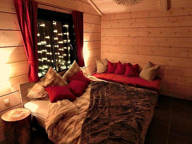 Schlafzimmer mit Holzwänden, Bett und Sofa mit Kissen. Fenster mit roten Vorhängen und Lichterkette.