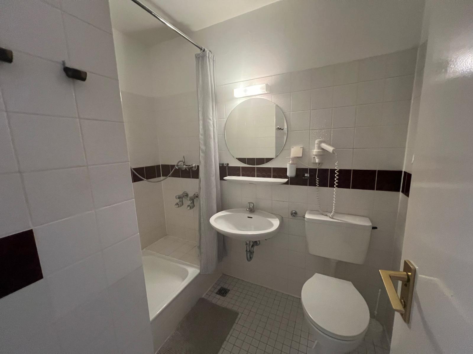 Badezimmer mit Dusche, Waschbecken und Toilette. Weiße Fliesen und runde Spiegel.